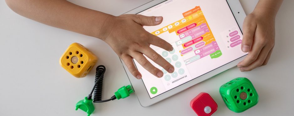 small hands on an white tablet navigating a mobile app with brightly colored toys and blocks on a white table